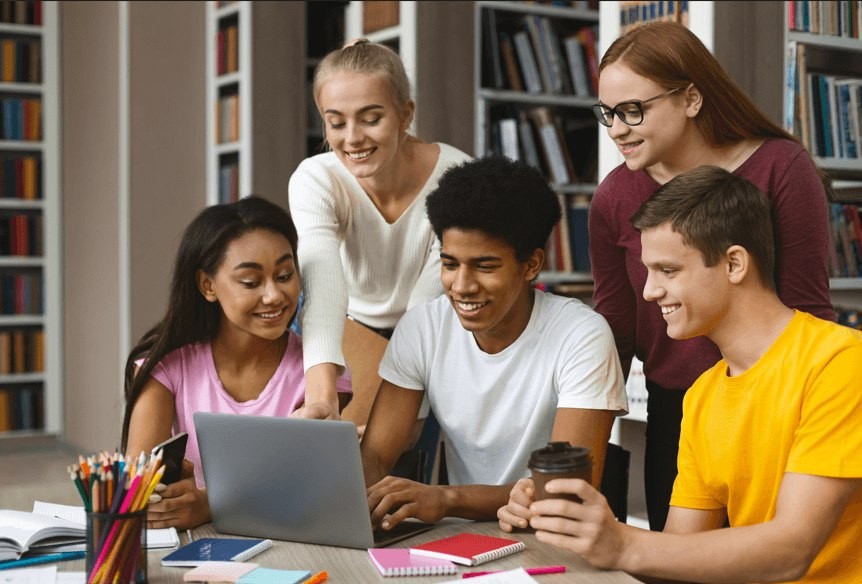 Group of five teenagers together in a library all looking at something on a laptop that is sitting on a table
